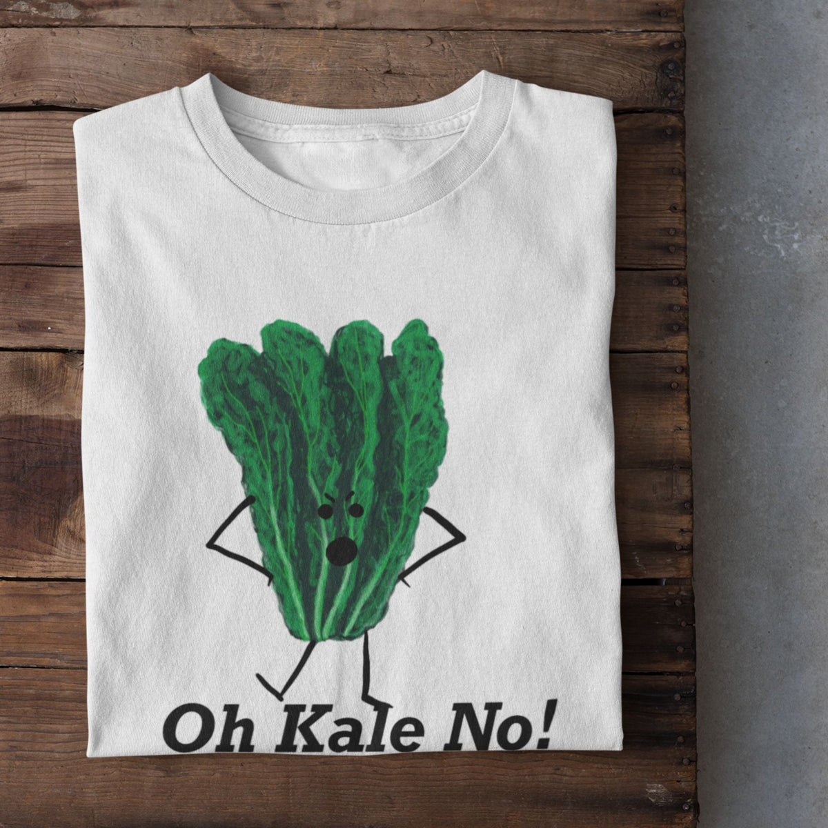 A folded white t-shirt with a green kale character graphic and the text 'Oh Kale No!' is displayed on a rustic wooden surface, with white sneakers partially visible at the bottom.