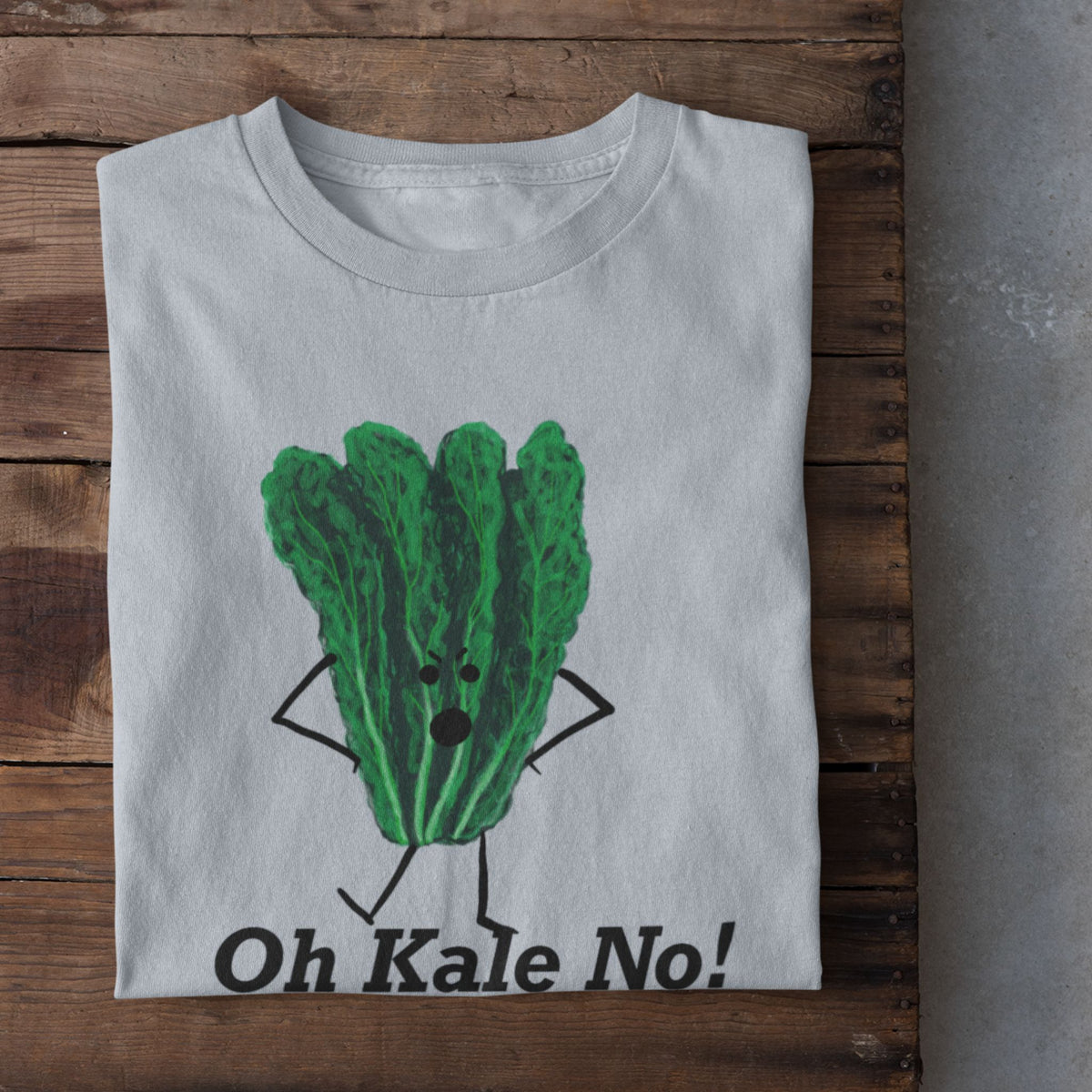 A light blue t-shirt laid flat on a rustic wooden crate, featuring a cartoon graphic of a green kale character with arms and legs, above the text 'Oh Kale No!'. White sneakers are visible at the bottom edge on a concrete floor.
