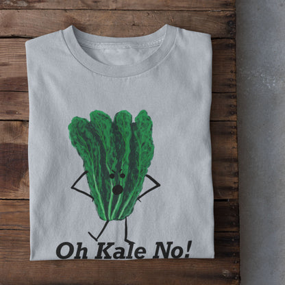 A light blue t-shirt laid flat on a rustic wooden crate, featuring a cartoon graphic of a green kale character with arms and legs, above the text 'Oh Kale No!'. White sneakers are visible at the bottom edge on a concrete floor.