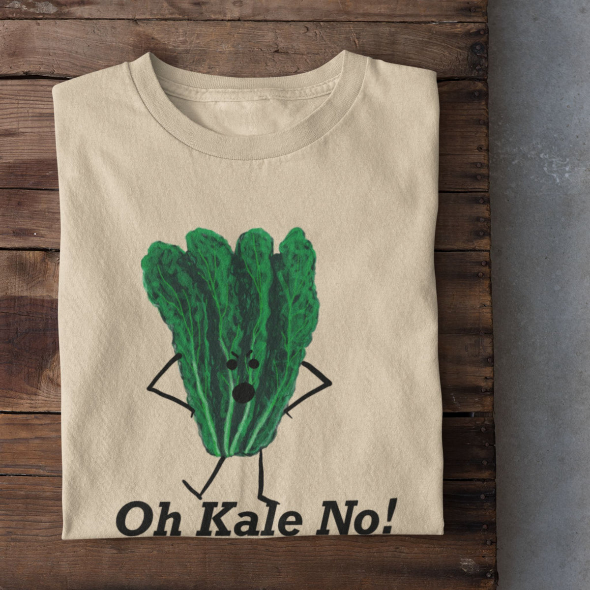 A soft cream crewneck t-shirt is laid flat on a rustic dark wood surface, featuring a cartoon graphic of a green kale vegetable with arms and legs, above the text 'Oh Kale No!'. White sneakers are visible at the bottom edge on a gray concrete floor.