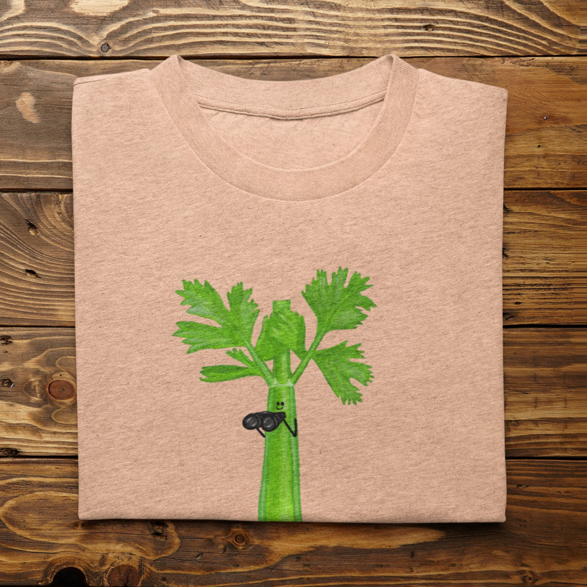 Heather peach Stalker celery pun T-shirt flat lay featuring a green celery character holding binoculars.
