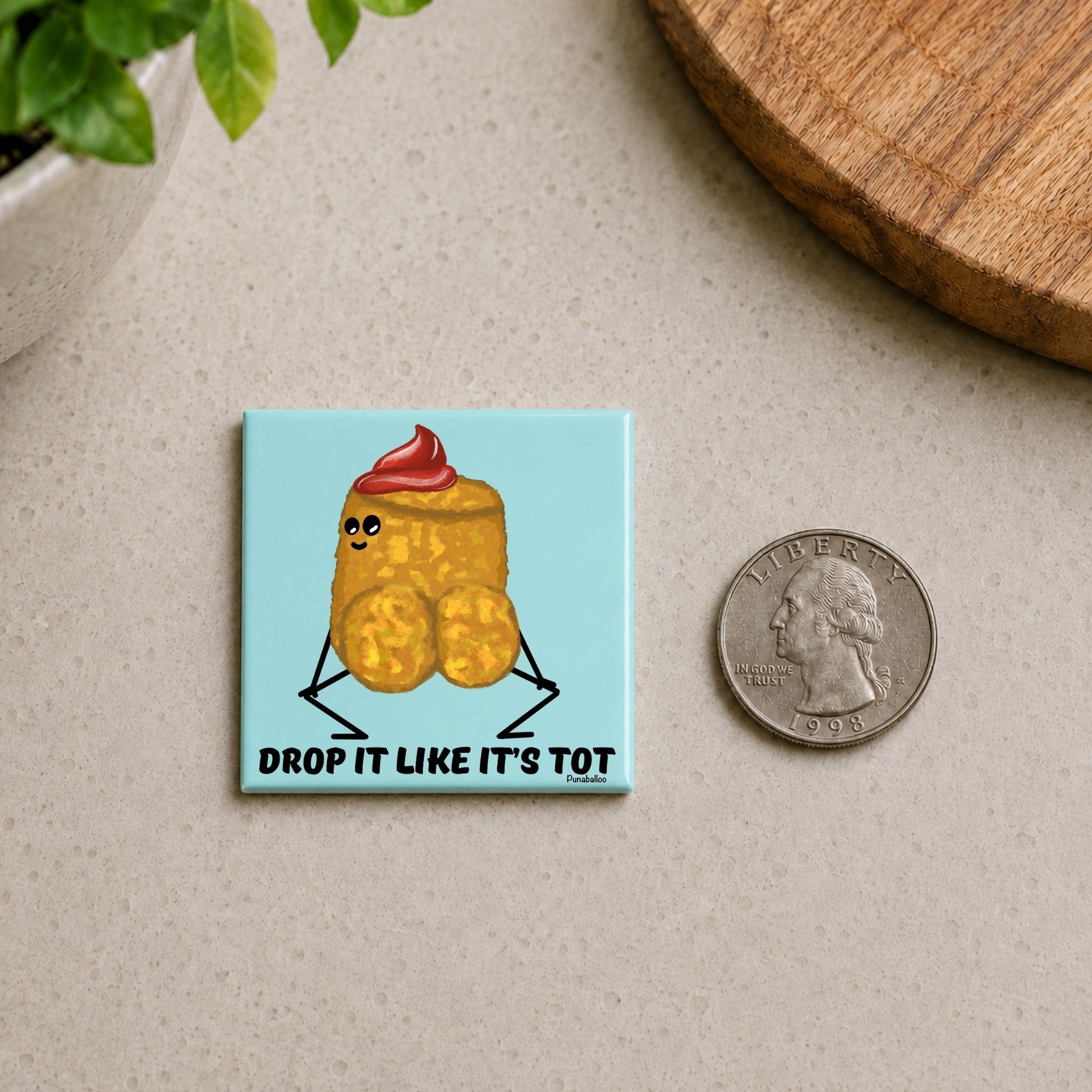 Drop It Like It’s Tot tater tot pun magnet on a countertop beside a quarter for size