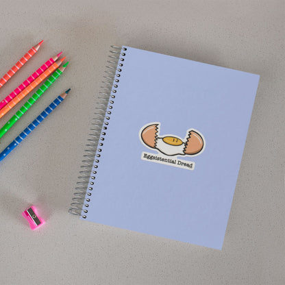 Eggsistential Dread dark humor food sticker on a notebook beside colored pencils