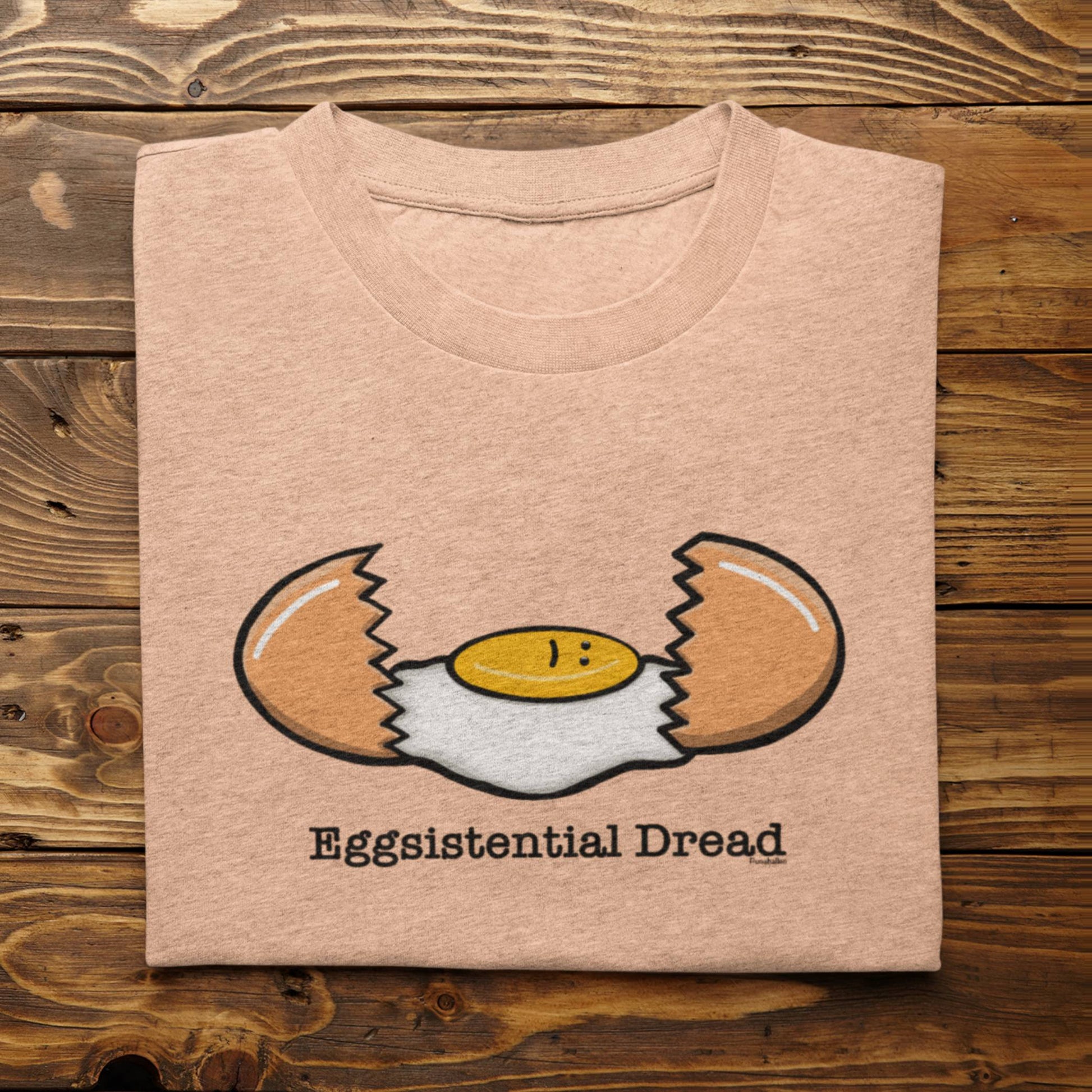 Folded heather peach Eggsistential Dread funny egg t-shirt on rustic wood background