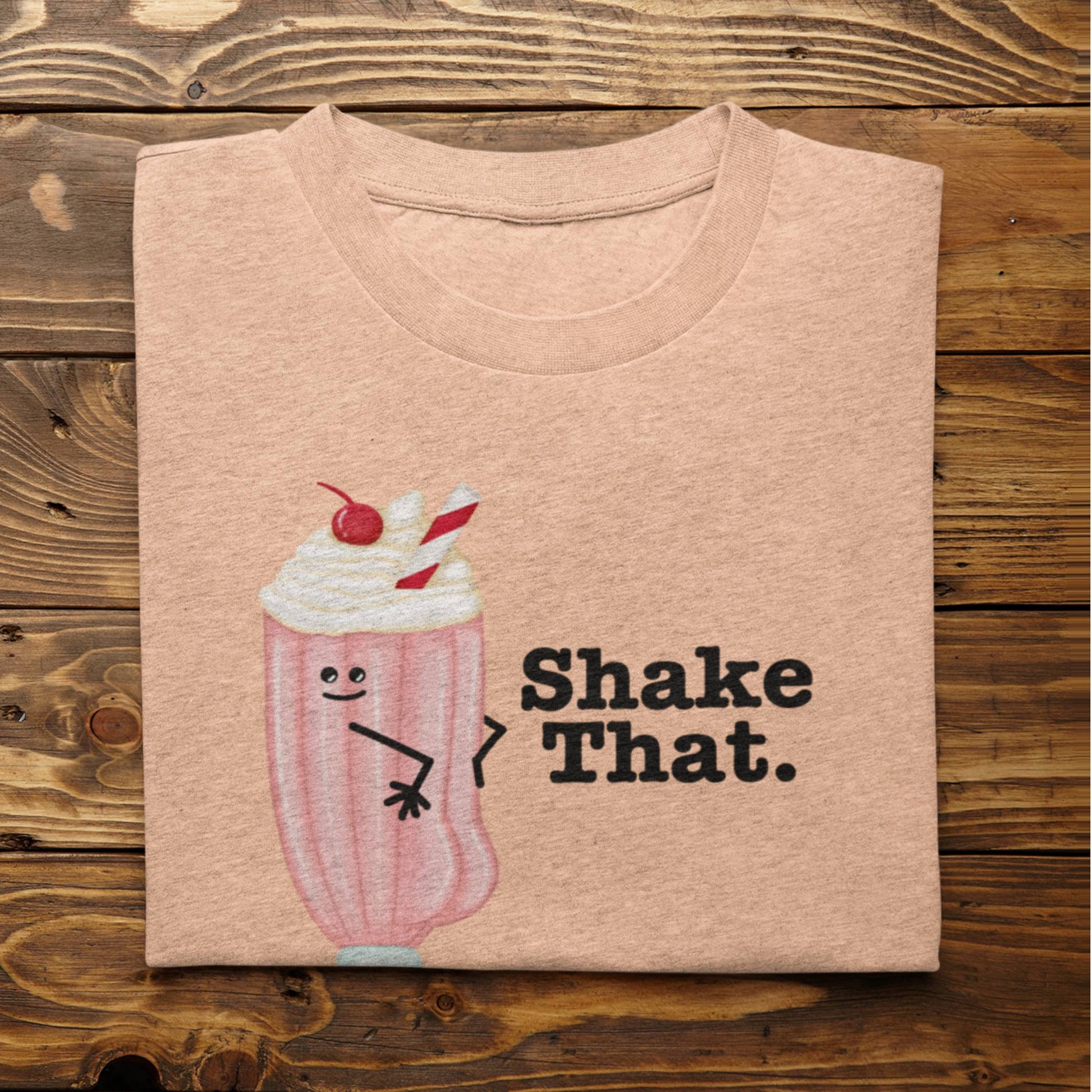 Folded heather peach Shake That milkshake pun t-shirt on rustic wood background