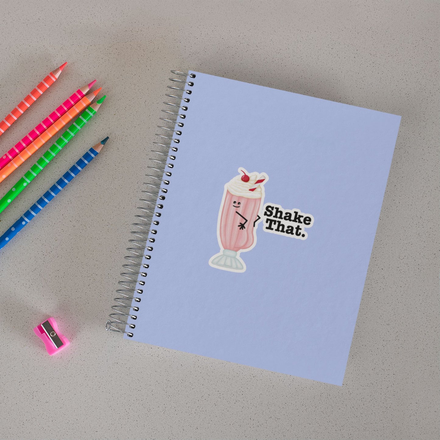 Shake That ice cream pun sticker on a notebook beside colored pencils