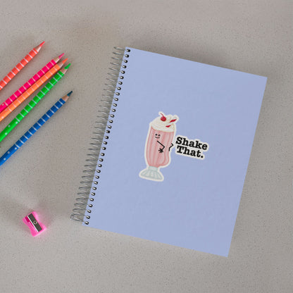 Shake That ice cream pun sticker on a notebook beside colored pencils