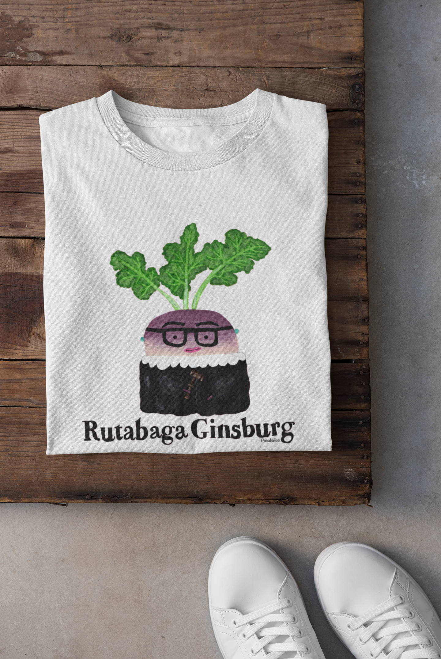 White t-shirt with a graphic design of a rutabaga wearing glasses and text 'Rutabaga Ginsburg' on a wooden surface.
