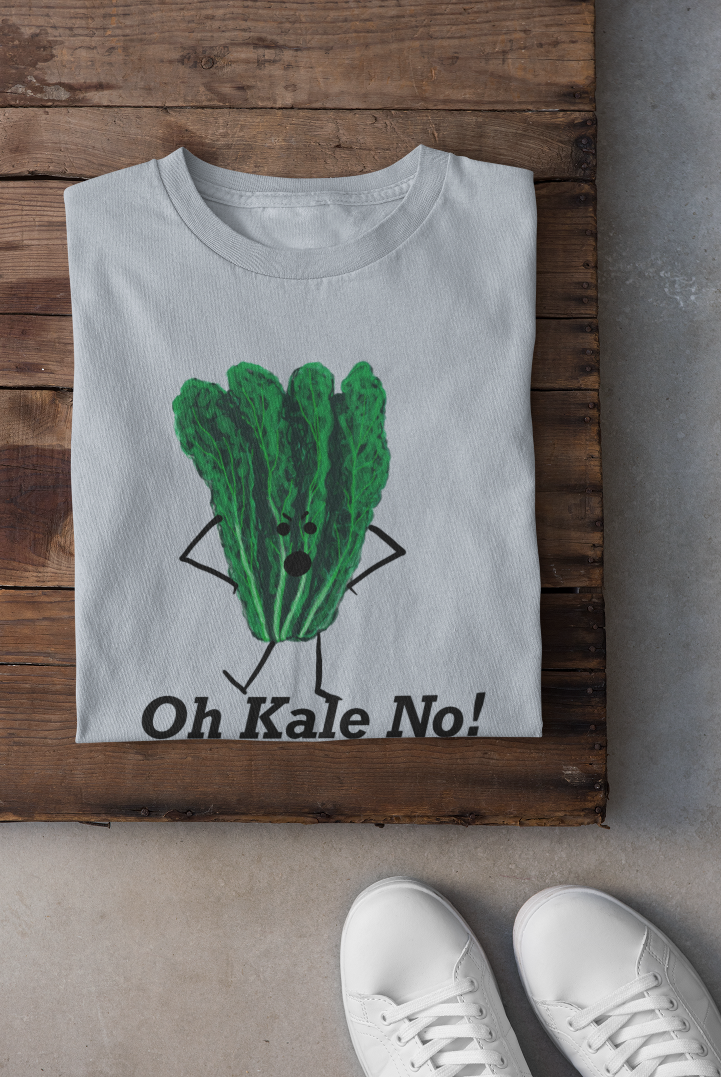 A light blue t-shirt laid flat on a rustic wooden crate, featuring a cartoon graphic of a green kale character with arms and legs, above the text 'Oh Kale No!'. White sneakers are visible at the bottom edge on a concrete floor.