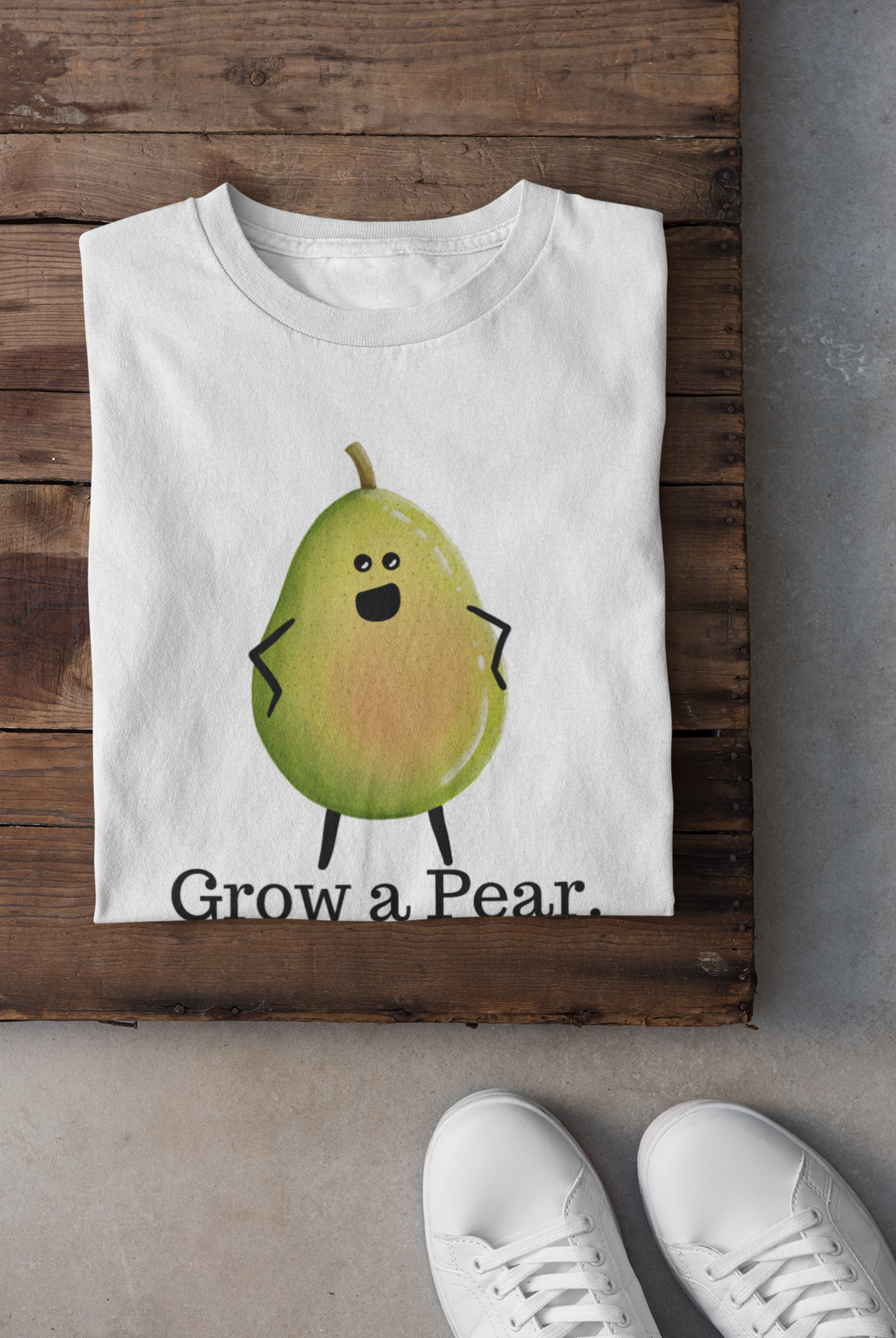 A folded white T-shirt with a cartoon pear character and the text 'Grow a Pear.' displayed on a rustic wooden surface, with white sneakers partially visible at the bottom right.