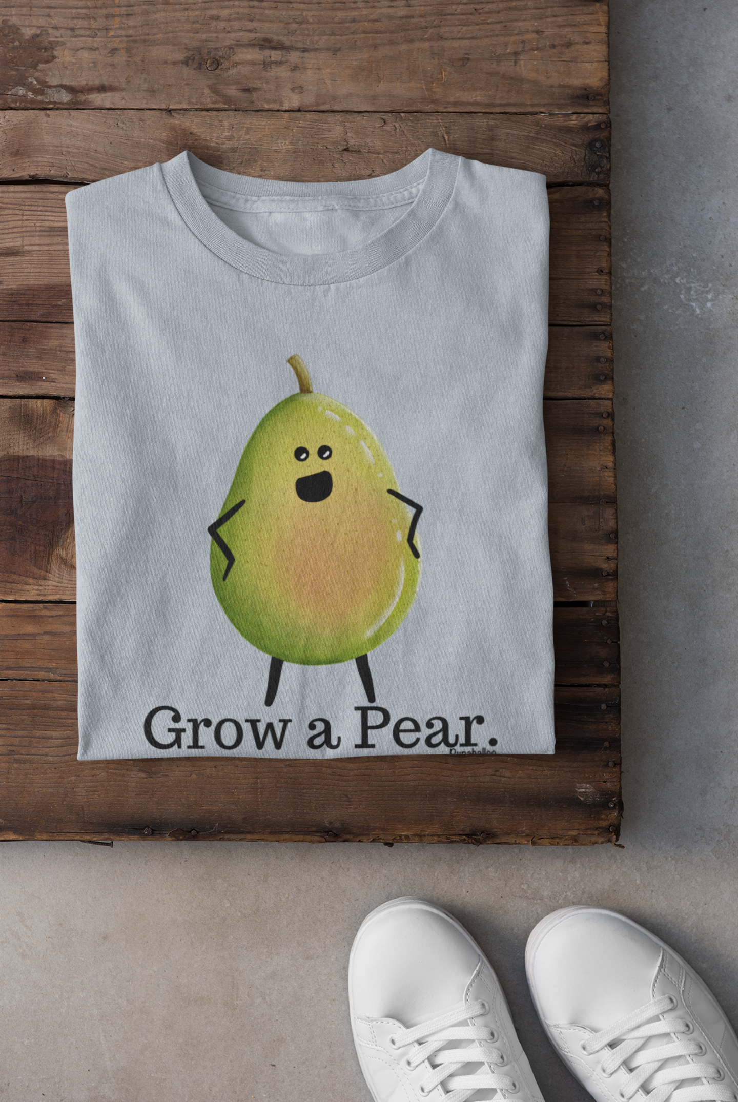 A folded light blue T-shirt with a cartoon pear character and the text 'Grow a Pear.' laid flat on a rustic wooden surface, with white sneakers partially visible at the bottom right.
