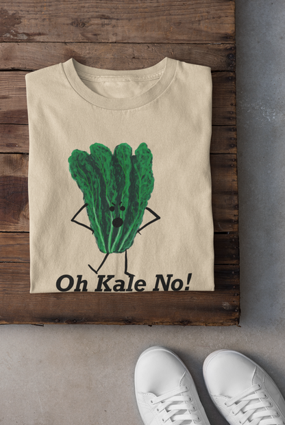 A soft cream crewneck t-shirt is laid flat on a rustic dark wood surface, featuring a cartoon graphic of a green kale vegetable with arms and legs, above the text 'Oh Kale No!'. White sneakers are visible at the bottom edge on a gray concrete floor.