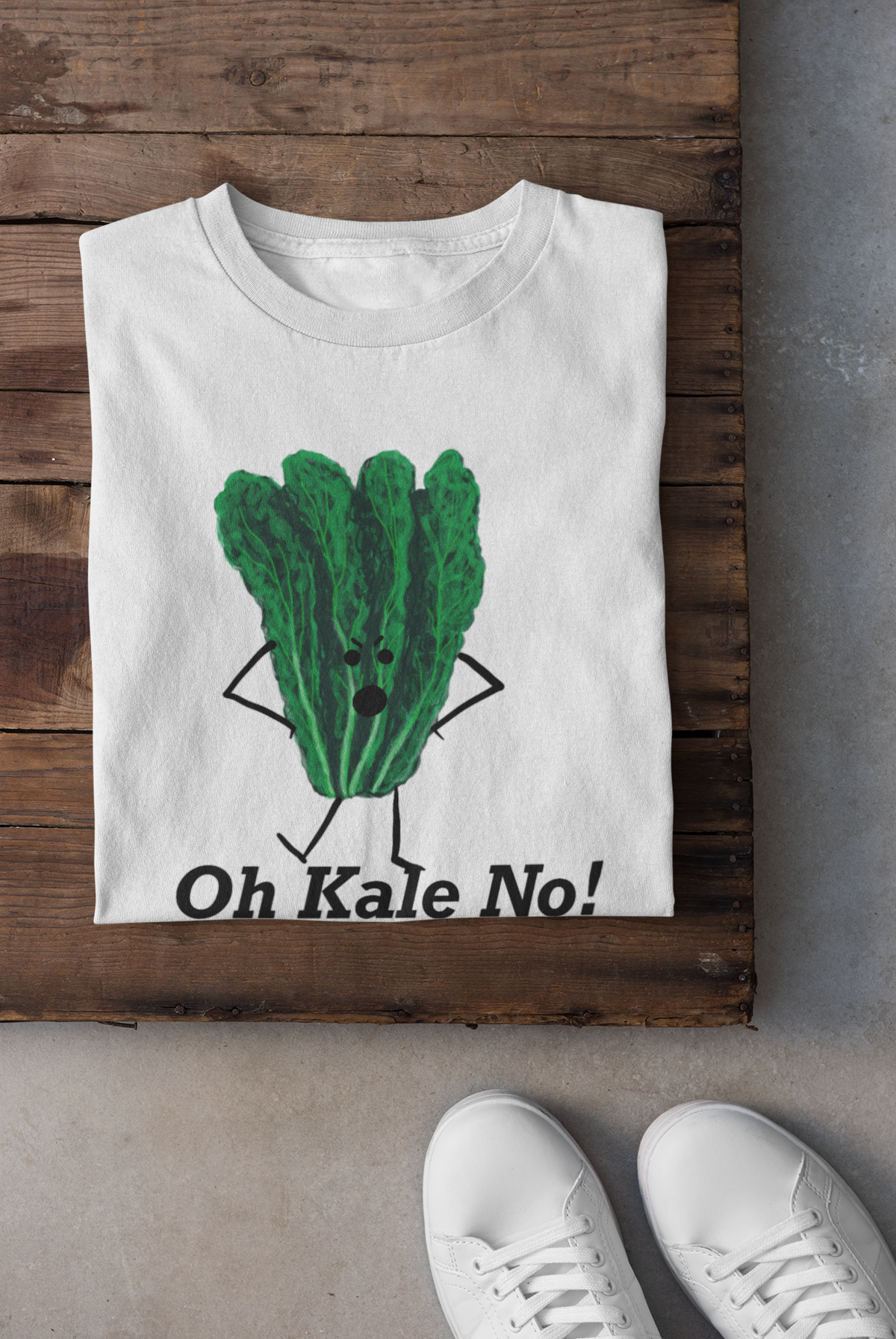 A folded white t-shirt with a green kale character graphic and the text 'Oh Kale No!' is displayed on a rustic wooden surface, with white sneakers partially visible at the bottom.
