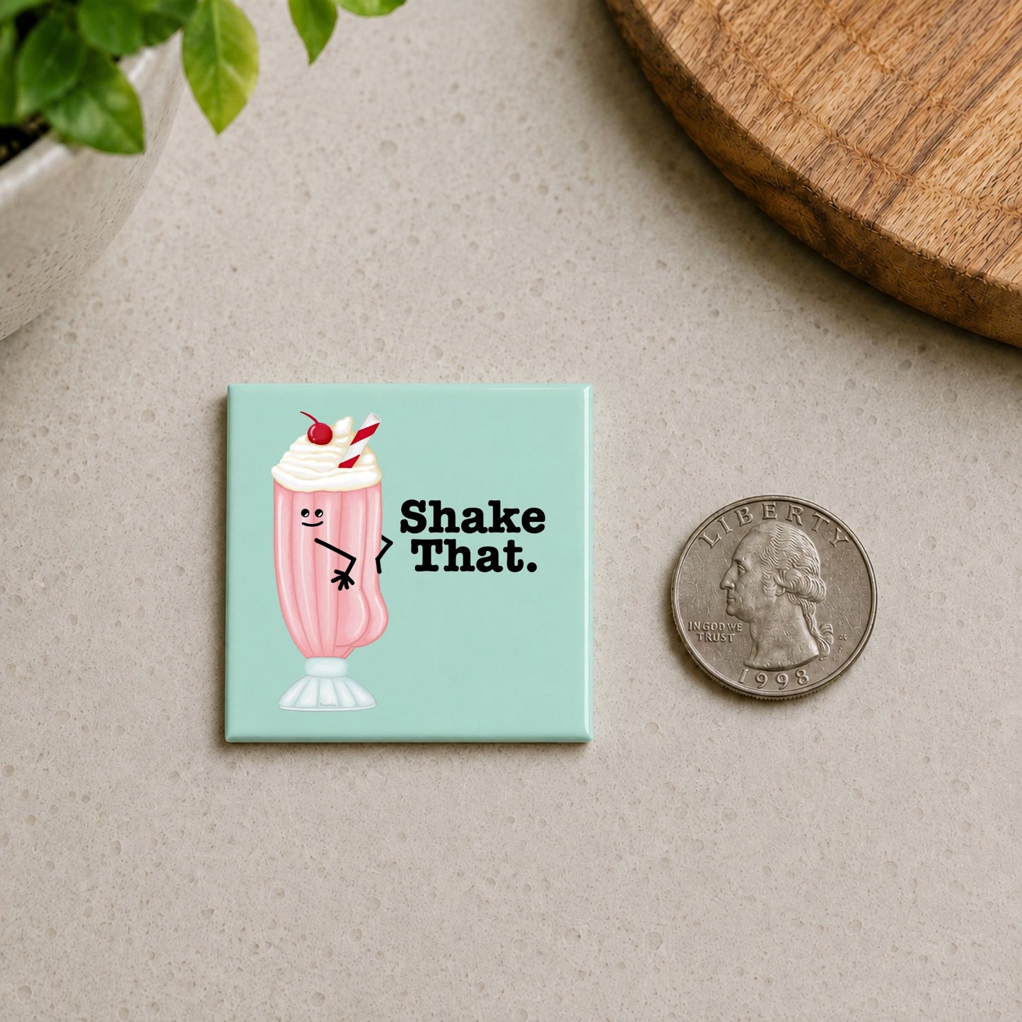 Shake That milkshake pun magnet shown beside a quarter for scale