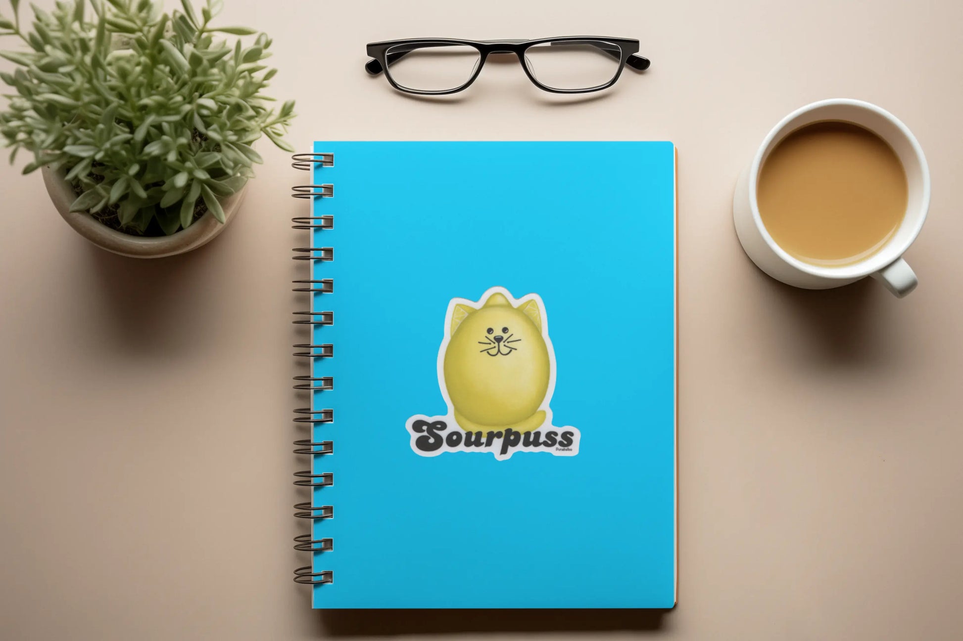 Blue notebook with a cartoon cat design and 'Sourpuss' text on a desk with a plant and coffee cup.