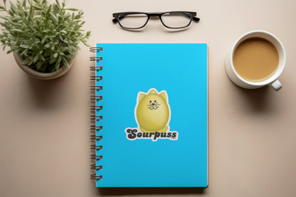 Blue notebook with a cartoon cat design and 'Sourpuss' text on a desk with a plant and coffee cup.