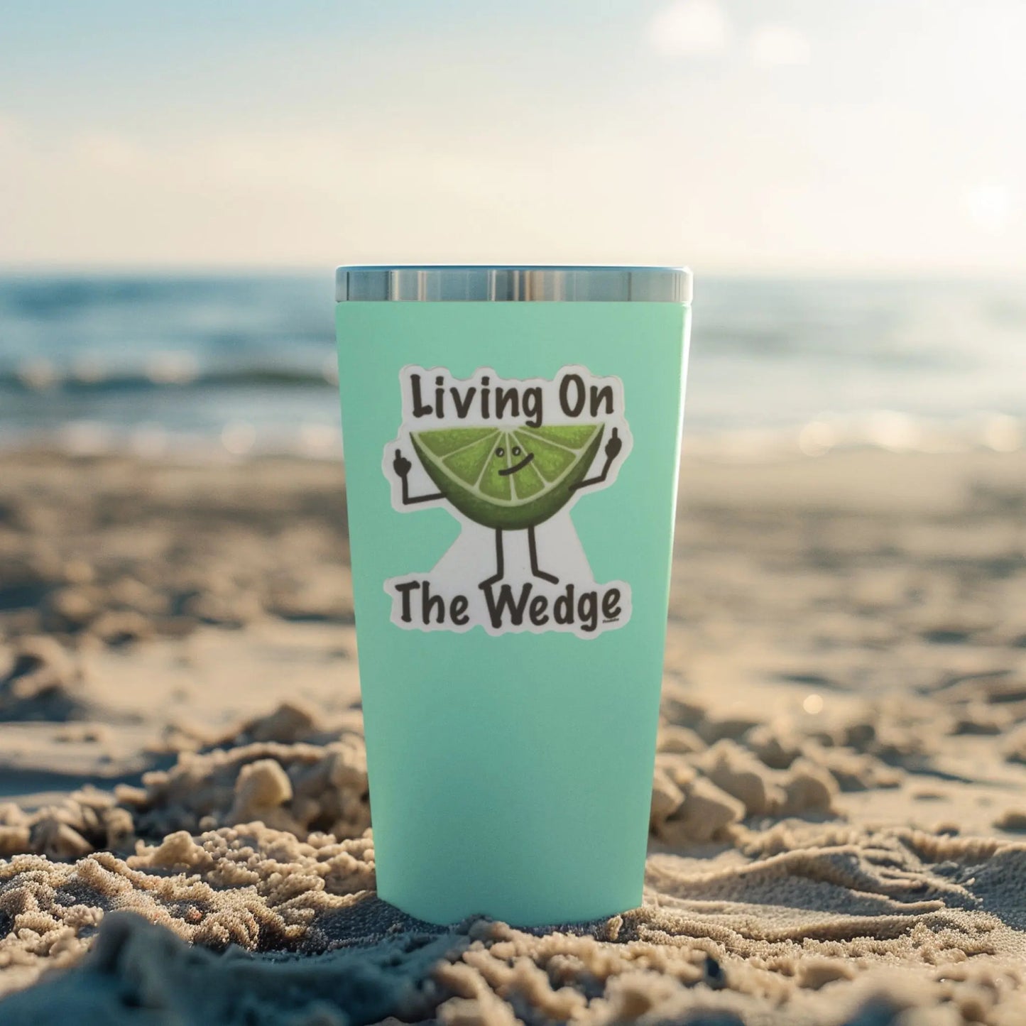 Turquoise tumbler with 'Living On The Wedge' fruit pun sticker on a sandy beach