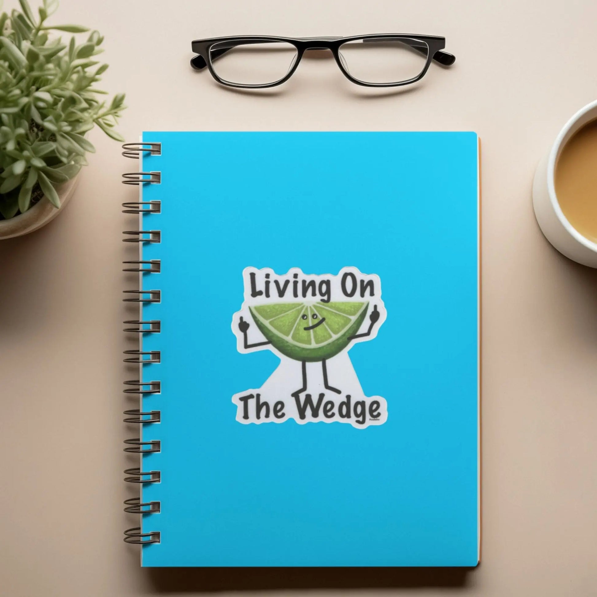 Blue notebook with 'Living On The Wedge' fruit sticker, glasses, plant, and coffee cup on a beige surface