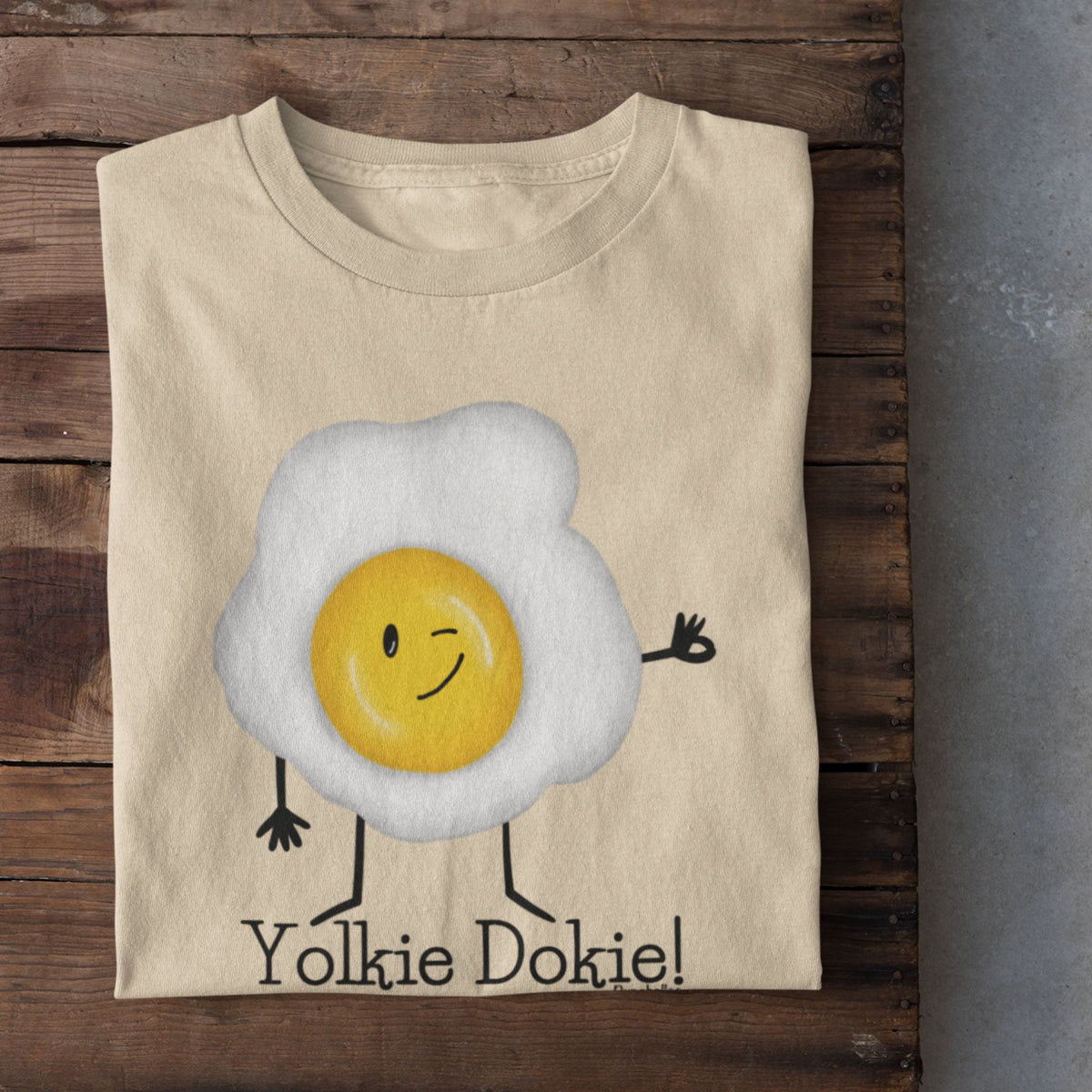 Soft Cream Yolkie Dokie fried egg pun T-shirt folded on a rustic wood background